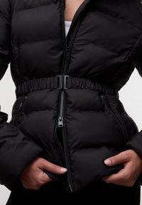 Zbliżenie na osobę ubraną w czarną pikowaną kurtkę z zamkiem błyskawicznym z przodu, elastycznym paskiem na klamrę oraz schowanymi w kieszeniach rękami.