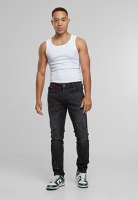 Een jonge man staat gekleed in een witte mouwloze shirt, zwarte ripped jeans en groen-witte sneakers, tegen een effen grijze achtergrond.