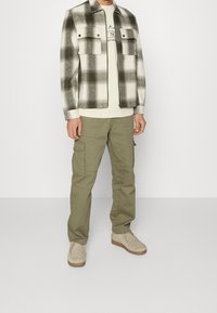 Chaqueta a cuadros en tonos gris y blanco con dos bolsillos en el pecho, combinada con pantalones cargo verde claro y zapatos de ante beige. Vista de cuerpo completo.