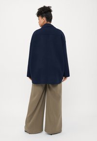 Námořnická oversized bunda s límcem, spadloumi rameny a dlouhými rukávy, v kombinaci s volnými khaki kalhotami, z pohledu zezadu.