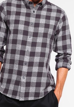 Mann trägt ein graues und schwarzes kariertes Hemd mit aufgekrempelten Ärmeln, eine Hand in der Tasche, vor einem schlichten weißen Hintergrund.