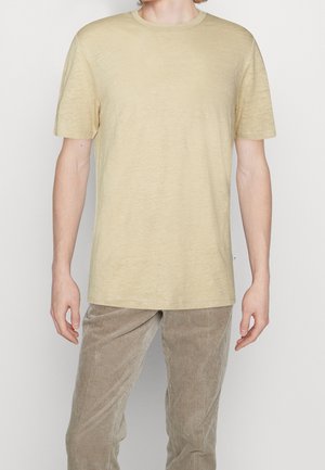 Mężczyzna w prostym beżowym t-shircie z krótkim rękawem i jasnobrązowych spodniach sztruksowych, stojący na białym tle.