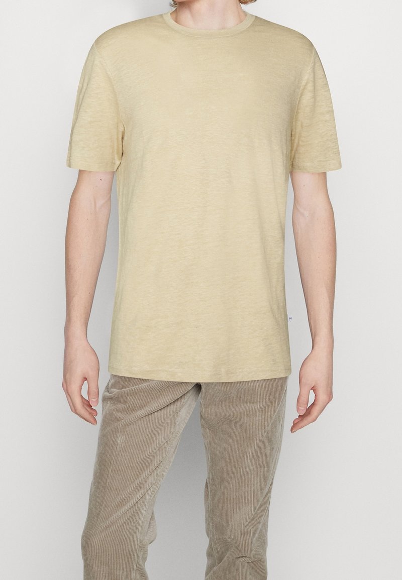 Homme portant un t-shirt beige uni à manches courtes et un pantalon en velours côtelé marron clair, debout devant un fond blanc.