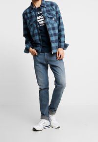Giacca a quadri blu e nera con bottoni, t-shirt blu Navy con testo bianco, jeans azzurri chiari e sneakers bianche con accenti navy.
