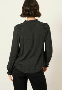 Frau mit kurzen braunen Haaren trägt eine schwarze Bluse mit langen Ärmeln, die weiße Punkte hat, und schwarze Hosen. Sie steht vor einer schlichten, hellen Wand.