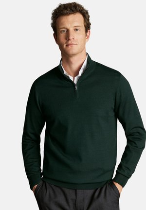 Man wearing dark green quarter-zip sweater over white shirt with hands in pockets, standing against plain light background.