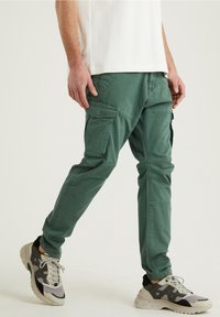 Cargo-byxor i mörkgrön bomull med flera sidofickor, avsmalnande ben och en matt yta, tillsammans med ljusa sneakers.