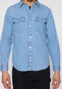 Camicia di denim azzurro chiaro con maniche lunghe, colletto a punta, due tasche sul petto e bottoni a pressione argentati; texture liscia e orlo curvo.