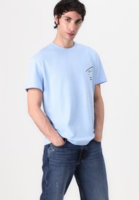 T-shirt en coton bleu clair avec des manches courtes, un col rond et un petit imprimé graphique noir sur la partie gauche de la poitrine.