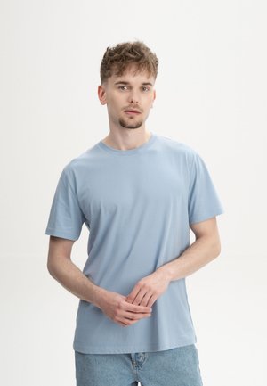 Jonge man met kort krullend haar draagt een lichtblauw T-shirt en spijkerbroek, staat met handen ontspannen gevouwen tegen een effen witte achtergrond.