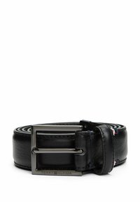 Zwarte leren riem met een zilverkleurige metalen gesp, een gladde textuur, zichtbare stiksels en een klein vlagaccent nabij de gesp.