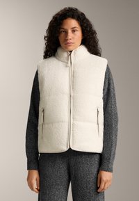 Frau trägt eine ärmellose cremefarbene Fleece-Weste mit Reißverschluss über einem dunkelgrauen, gestrickten Langarmshirt und Hose, steht vor einem neutralen Hintergrund.