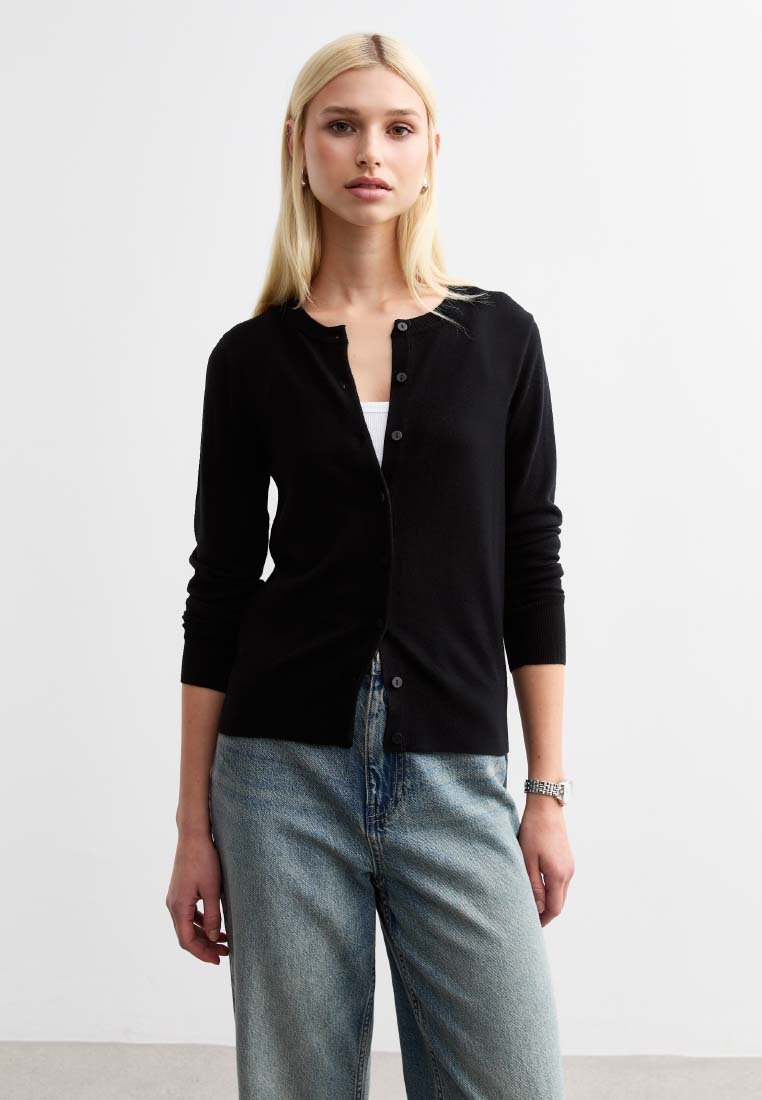 Cardigan nero con scollatura tonda, maniche lunghe e chiusura con bottoni. Indossato sopra una maglietta bianca, abbinato a jeans azzurri chiari.