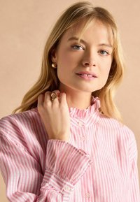 Blonde woman wearing a pink and white striped blouse with a ruffled collar, holding her hand near her neck, wearing gold earrings and rings.