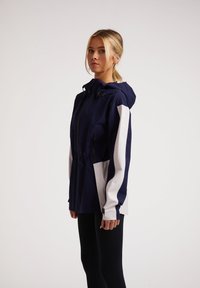 Chaqueta impermeable en navy y blanca con capucha, que cuenta con bolsillos con cremallera, mangas en contraste y un corte relajado para mayor libertad de movimiento.