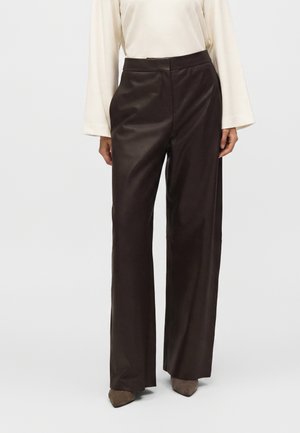 Donna che indossa pantaloni in pelle a vita alta, a gamba larga, marrone scuro, con scarpe beige con punta e un top a manica lunga color avorio.
