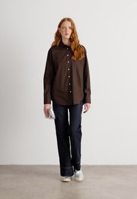Brown button-up shirt with a pocket, paired with dark denim flared jeans and silver sneakers. The figure holds a clear clutch.