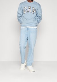 Felpa blu con logo "GAP" in crema sul petto, abbinata a jeans azzurri chiari e sneakers bianche. Tessuto morbido e vestibilità comoda.