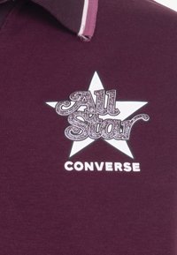 Maroon majica s kragnom s bijelim grafičkim uzorkom zvijezde, s natpisom "All Star" u sjajnoj srebrnoj boji i "CONVERSE" ispisanim u podebljanoj bijeloj boji ispod.