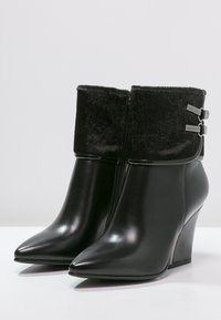 Botines de cuero negro con un puño de piel texturizada, punta afilada y tacón en cuña. Presentan hebillas plateadas y una cremallera elegante.