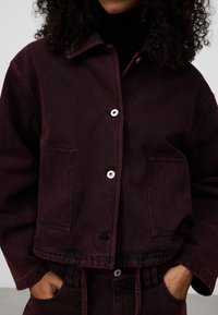 Burgunderfärgad jeansjacka med krage, två framfickor och knäppning. Har subtil textur och kontrasterande sömmar.