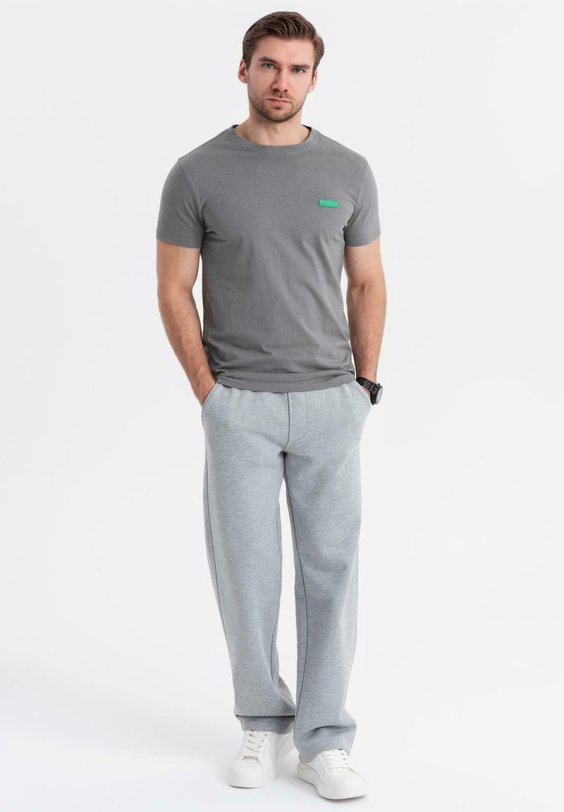 T-shirt grigio a maniche corte con logo verde, abbinato a pantaloni della tuta grigi chiari e sneakers bianche, creando un outfit casual.