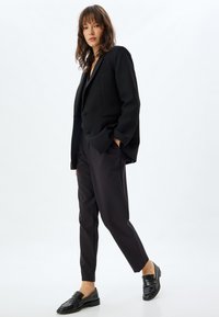Blazer negro de gran tamaño con dos botones, combinado con pantalones negros a medida. Mocasines de cuero negro con detalle texturizado. Fondo liso.