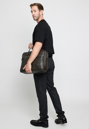 Man wearing black t-shirt and dark pants holding a dark leather briefcase, standing against a plain light gray background.