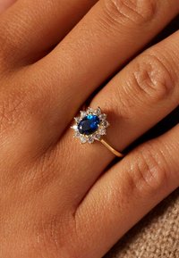 Gold ring featuring an oval blue gemstone at the center, surrounded by a halo of small clear stones, set on a textured hand.