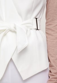 Biała kamizelka z przewiązaniem w talii i metalową klamrą, o gładkiej fakturze. Pod spodem częściowo widoczna jest jasnoróżowa koszula z długim rękawem.