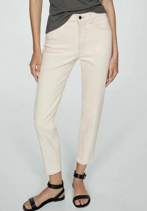 Vrouw draagt off-white cropped broek, zwarte sandaaltjes met enkelriem en een ingestopt donkergrijs topje, staat met armen ontspannen langs het lichaam.