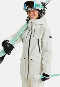 Weiße Skijacke mit schwarzen Reißverschlüssen, Taschen und Kapuze. Schwarze Handschuhe, die mintfarbene Skier halten. Skibrille auf dem Helm.