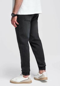 Joggers negros ajustados con puños elásticos, combinados con zapatillas blancas y beige. Incluyen textura de denim suave y sutiles detalles de bolsillo.