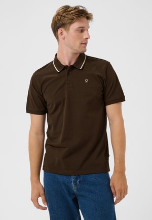 Hombre con camiseta polo marrón de manga corta con raya blanca en el cuello y vaqueros azul, mirando hacia su izquierda sobre un fondo liso.