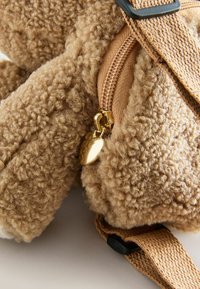 Tan, fluffy materiaal tas met een gestructureerde oppervlakte en een gouden ritsaccent. Bevat een hartvormige charme; verstelbare schouderband zichtbaar.