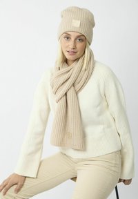 Ensemble bonnet et écharpe en tricot beige présenté sur un modèle, avec des motifs texturés, un patch logo, et un pull et pantalon de couleur crème.