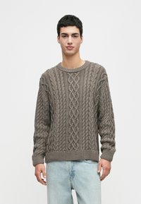 Junger Mann, der einen grauen Zopfmustersweater und hellblaue Jeans trägt, steht vor einem schlichten weißen Hintergrund.