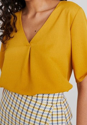 Woman wearing a mustard yellow V-neck blouse with short sleeves and a high-waisted skirt featuring a yellow, blue, black, and white plaid pattern.