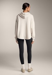 Hanorac oversize de culoare bej deschis, cu glugă, asortat cu colanți negri și șosete scurte de culoare crem. Textură moale și croială lejeră.