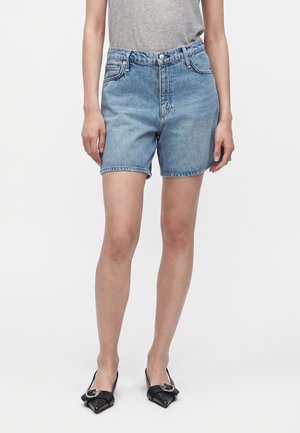 Personne portant un short en jean bleu clair, un t-shirt gris rentré et des ballerines noires pointues avec des détails de boucle, debout devant un fond uni.