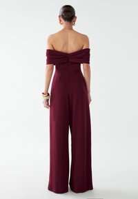 Bordeauxroter Schulterfreier Jumpsuit mit tailliertem Oberteil, weit geschnittenen Hosenbeinen und Reißverschluss am Rücken. Glatte Textur mit drapierten Ärmeln.