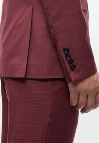 Burgundi színű öltöny texturált anyagból. A zubbon ujjának közeli felvétele négy fekete gombot mutat, összehangolt cérna részletekkel és redővel.