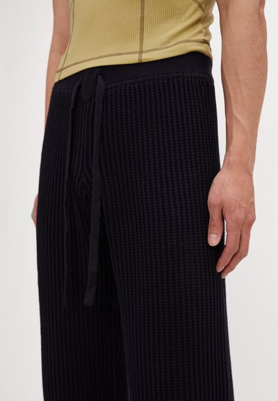Pantaloni tricotati texturati de culoare bleumarin închis, cu talie elastică și sistem de închidere cu șnur. Prim-plan al țesăturii și al unei mâini așezate pe picior.
