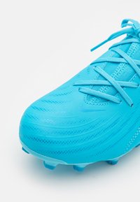 Turkosfärgad fotbollssko med texturerad syntetisk ovandel, förstärkt tå och platta snören. Detaljerna inkluderar en elegant design och en böjd form.