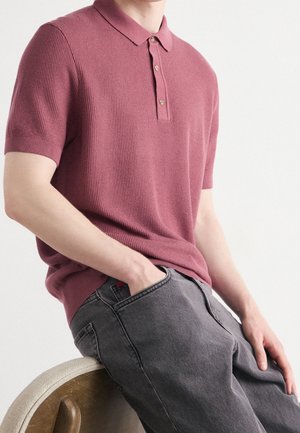 Personne portant un polo à manches courtes mauve texturé et un jean gris foncé, assise sur une chaise avec une main dans la poche.