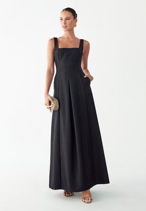 Woman wearing a sleeveless black floor-length dress, holding a beige clutch, with gold earrings, bracelet, and sandals on a white background.