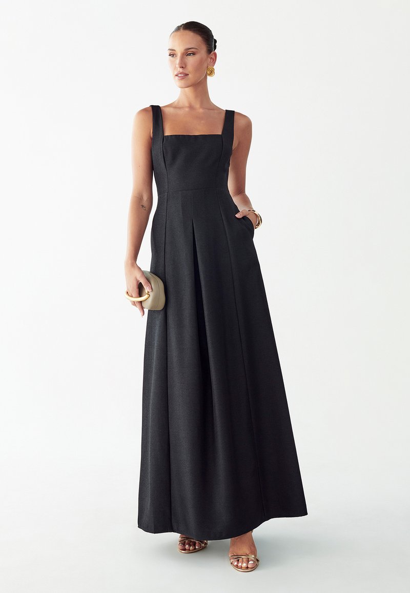 Žena v čiernych dlhých šatách bez rukávov, drží béžové psaníčko, so zlatými náušnicami, náramkom a sandálmi na bielom pozadí.