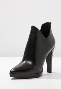 Botín de tacón alto de cuero negro con puntera afilada, corte lateral y tacón delgado apilado sobre una superficie blanca.