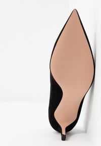 Chaussure à talon kitten en daim noir avec bout pointu, positionnée debout montrant une semelle beige lisse sur fond blanc.