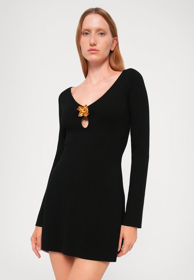Black ribbed dress with long sleeves, featuring a deep V-neckline and a gold floral brooch accent at the center. Fitted silhouette.